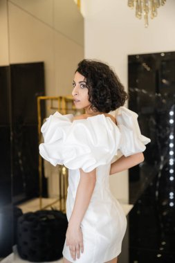Ortadoğulu, esmer, dalgalı saçlı, kabarık kollu ve fırfırlı moda gelinlik deneyen ve modern gelin butiği, yansıması, alışverişi, müstakbel gelini, zarif bakışları olan bir kadın. 