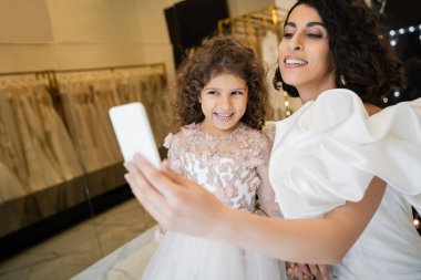 Çekici bir ortadoğulu gelin, esmer saçlı, beyaz gelinlikli, kabarık kollu, fırfırlı elbiseli, akıllı telefondan selfie çeken mutlu gelinlik mağazasında geçmişi bulanık, mutlu bir kızla birlikte. 
