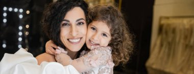 Çiçek gibi giyinmiş neşeli küçük bir kız beyaz gelinlikli, kabarık kollu ve fırfırlı güzel annesini sıkıca kucaklarken gelin butiğinde birlikte kameraya bakıp gülümsüyor. 