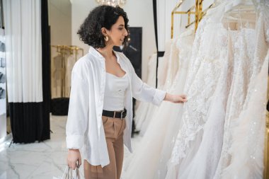 Kahverengi saçlı, dalgalı saçlı, beyaz pantolonlu, elinde alışveriş torbalarıyla gelin butiğinde gelinlik seçen mutlu orta doğulu kadının yan görüntüsü. 