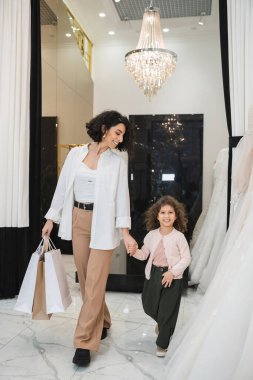 Kahverengi pantolonlu, beyaz gömlekli, alışveriş çantalı mutlu bir orta doğulu kadın gelinlik salonundaki gelinliğin yanında neşeli kızıyla yürürken, modern gelin, anne...