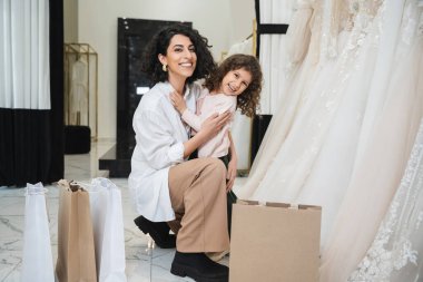 Küçük mutlu bir kız, esmer saçlı, beyaz gömlekli, neşeli bir ortadoğulu geline sarılıyor alışveriş çantaları ve gelinlik dükkanında oturuyor, anne-kız kaynaşıyor. 