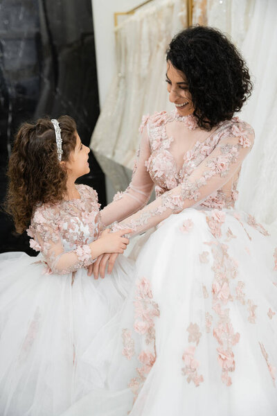 brunette middle eastern woman with wavy hair looking at girl and smiling near white wedding dresses in bridal salon, floral, mother and daughter, happiness, wedding day, shopping, bonding 