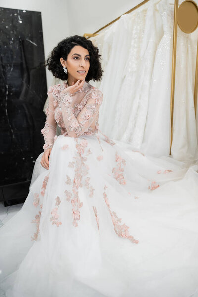 happy middle eastern woman with wavy hair sitting in gorgeous and floral wedding dress near blurred and white gown inside of luxurious bridal salon, charming and elegant, bride-to-be