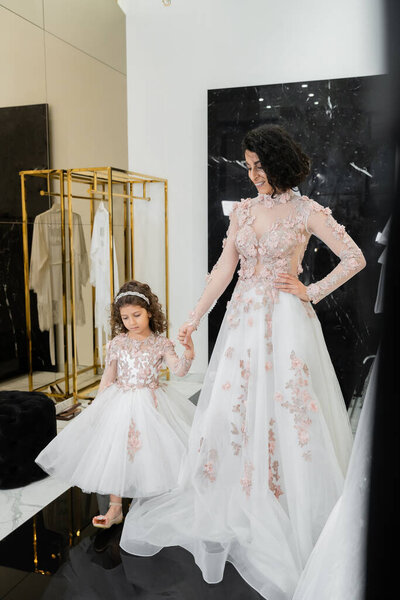 happy middle eastern woman with brunette wavy hair in stunning wedding dress holding hands with daughter in cute floral attire while standing in bridal salon, shopping, golden accents
