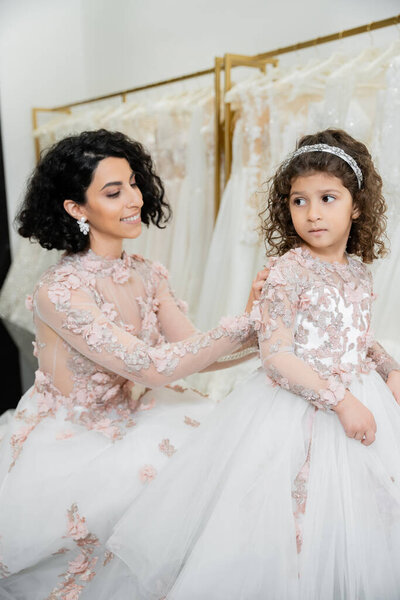 charming middle eastern bride with brunette wavy hair in wedding dress adjusting cute floral dress of daughter in bridal salon, shopping, special moment, togetherness, blurred white gown 