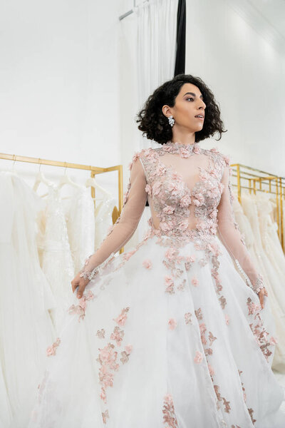 очаровательная и средневосточная женщина примеряет роскошное и цветочное свадебное платье внутри роскошного свадебного салона, делает покупки, невеста-будущая, размытое белое платье на заднем плане, отворачивается 