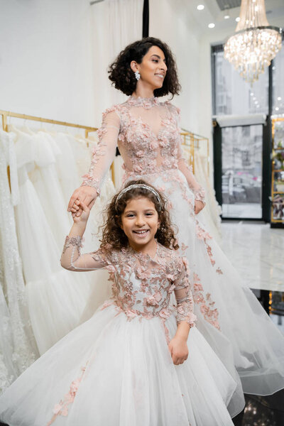 middle eastern girl in floral attire holding hands with cheerful woman standing in wedding dress near blurred white gown inside of luxurious bridal salon, shopping, bride-to-be, mother and daughter 