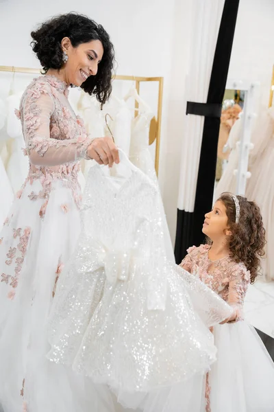 幸せとブルネット真ん中東部の花嫁で花の結婚式のガウンを選択するのを助けるドレスのために彼女の小さな娘のためのブライダルサロンで白いチュール生地の周り 準備のプロセス — ストック写真