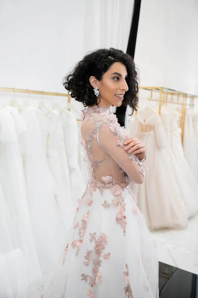 stock image cheerful middle eastern and brunette woman with wavy hair standing in gorgeous and floral wedding dress inside of luxurious bridal salon around white tulle fabrics, bridal shopping, looking away 