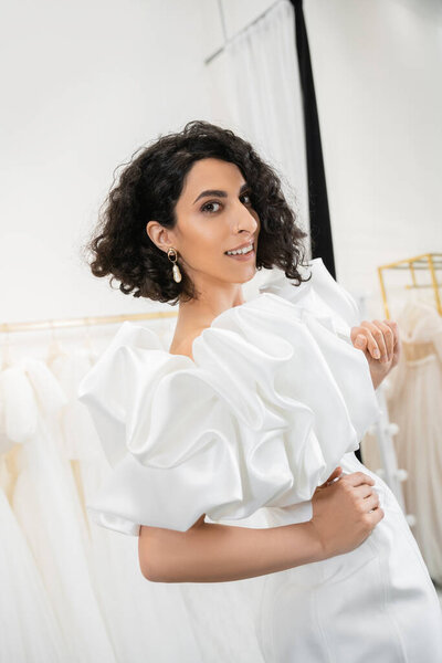 happy middle eastern bride with brunette and wavy hair posing with hands on hips in trendy wedding dress with puff sleeves and ruffles in bridal boutique next to tulle fabrics 