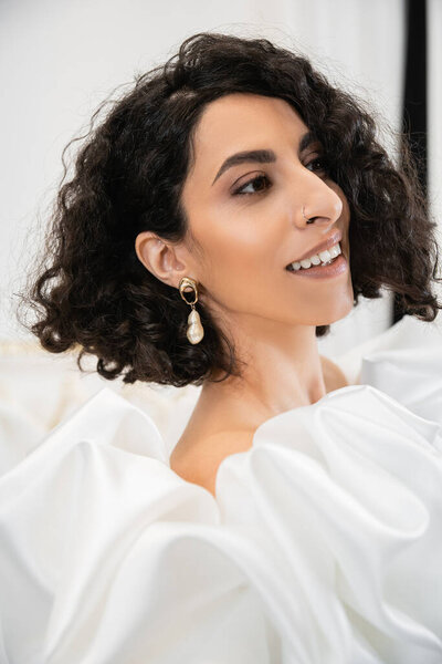 portrait of happy brunette middle eastern bride with wavy hair posing in trendy wedding dress with puff sleeves and ruffles in bridal boutique and looking away, delightful woman 