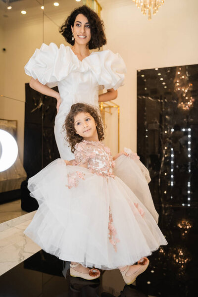 charming middle eastern bride with brunette hair standing in white wedding gown with puff sleeves and ruffles and looking at mirror near daughter holding tulle skirt in bridal store 