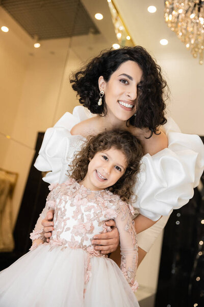 charming middle eastern bride with brunette hair standing in white wedding gown with puff sleeves and ruffles behind cute daughter and smiling together in bridal store, looking at camera 