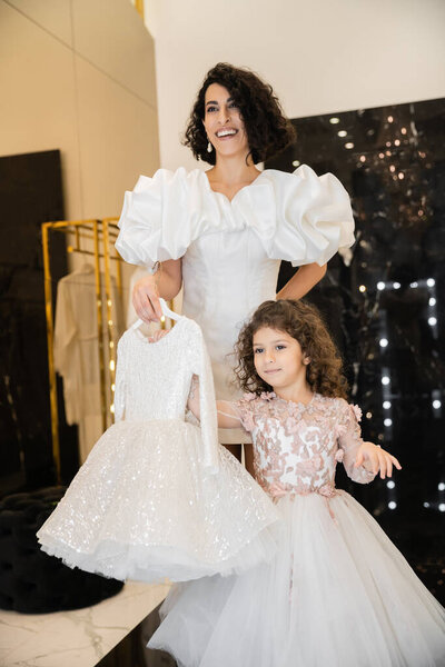 charming middle eastern bride with brunette hair standing in white wedding gown with puff sleeves and ruffles and holding girly dress with tulle skirt near daughter in bridal store 