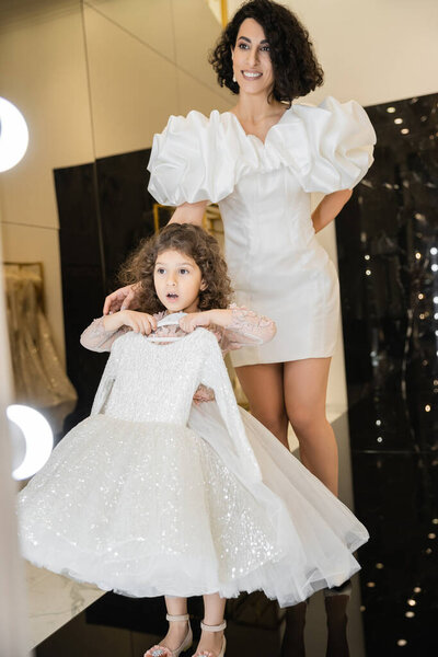 happy middle eastern bride with brunette hair standing in white wedding gown with puff sleeves and ruffles near surprised daughter holding girly dress with tulle skirt near mirror in boutique 