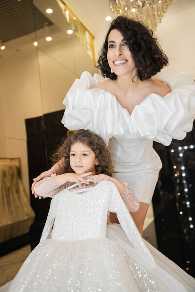 joyous middle eastern bride with brunette hair standing in white wedding gown with puff sleeves and ruffles near cute daughter holding girly dress with tulle skirt in bridal boutique  
