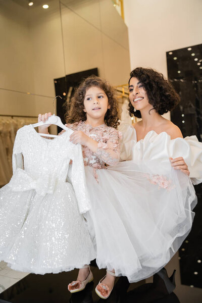 charming middle eastern bride with brunette hair standing in white wedding gown with puff sleeves and ruffles holding tulle skirt of her cute daughter holding girly dress in bridal boutique  