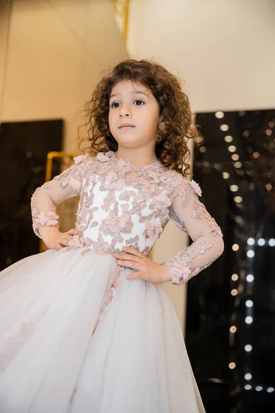 stock image curly middle eastern and little girl in floral dress with tulle skirt standing with hands on hips and looking away in bridal boutique, preparation for wedding, blurred background, golden accents