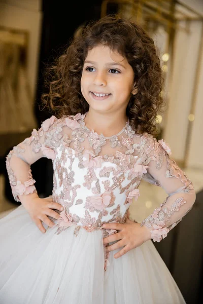 stock image joyous middle eastern and little girl in floral dress with tulle skirt standing with hands on hips and looking away in bridal boutique, preparation for wedding, blurred background 
