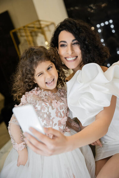 Очаровательная невеста с брюнетками в белом свадебном платье с пышными рукавами и оборками, делающая селфи на смартфоне со смешной дочерью, подмигивающей в свадебном магазине с размытым фоном 