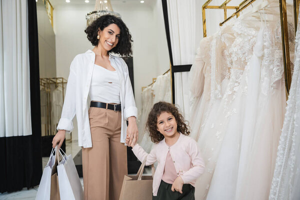 cheerful middle eastern bride with brunette hair in beige pants with white shirt holding shopping bags while standing with little girl near wedding dresses in bridal salon, mother and daughter 