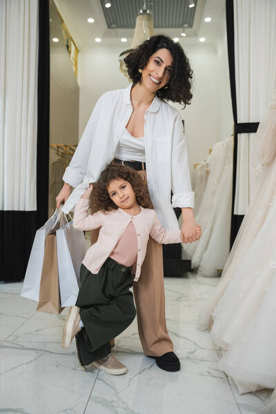 happy middle eastern woman with brunette hair holding shopping bags and hands of cute little girl while standing near wedding dresses in bridal salon, mother and daughter, bridal shopping 