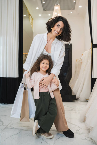 cheerful middle eastern woman with brunette hair holding shopping bags and hugging cute little girl while standing near wedding dresses in bridal salon, mother and daughter, bridal shopping 