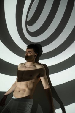 young, sexy and tempting man with shirtless body, muscular torso, in underpants posing while standing on abstract background with black and white spiral projection clipart