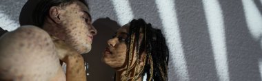 Çekici ve baştan çıkarıcı Afro-Amerikan kadınının düşük açılı görüntüsü şık rastalı, beyaz desenli arka planda beyaz gömleksiz, gri gölgeli, afişi olan seksi bir adamın yanında duruyor.