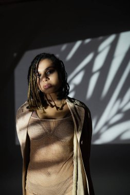 Çekici ve seksi Afro-Amerikalı kadın iç çamaşırlı, takım elbiseli ve ceketli siyah beyaz arka planda beyaz gölgelerle poz verirken kameraya bakıyor.