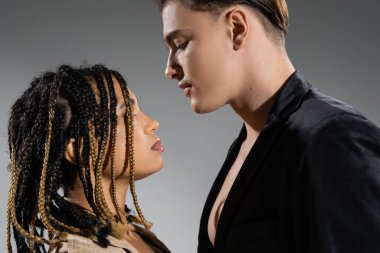 side view of sexy interracial couple, african american woman with stylish dreadlocks and young good looking man wearing black silk blazer on grey background clipart