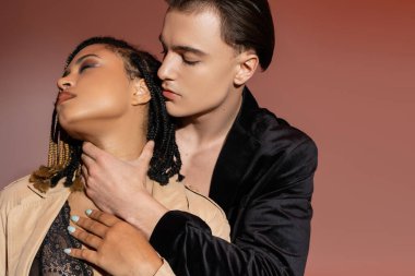 Siyah ipek ceketli genç ve şık bir adam dantelli iç çamaşırı ve pembe bej arka planda trençkot giyen baştan çıkarıcı Afrikalı Amerikalı bir kadının boynunu kucaklıyor.