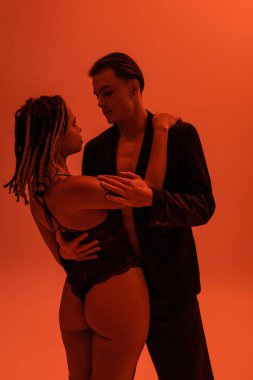 Siyah ceketli genç ve şık bir adam rastalı ve seksi kalçalı Afro-Amerikan kadınına sarılıyor turuncu arka planda dantel bir elbise giyiyor ve kırmızı ışık efekti kullanıyor.