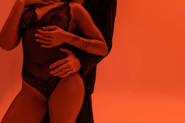 Siyah ceketli genç bir adamın, kollarında dövme olan, turuncu arka planda dantel ceket giyen seksi ve tutkulu Afro-Amerikan kadını kucaklaması.