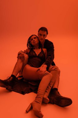 full length of young handsome man in blazer embracing sexy african american woman in black lace bodysuit and over knee boots while sitting on clothes on orange background with red lighting effect clipart