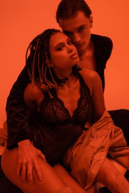 confident and sexy man with young and seductive african american woman in black lace bodysuit looking at camera while sitting on orange background with red lighting effect clipart