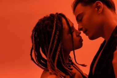 Genç ve şehvetli ırklar arası bir çiftin yan görünüşü, karizmatik bir adam ve turuncu arka planda kırmızı ışık efektiyle gözleri kapalı öpüşen Afro-Amerikan kadını.