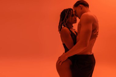 side view of young shirtless man with muscular torso hugging sexy african american woman with dreadlocks, wearing black lace bodysuit on orange background with red lighting effect clipart