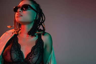 portrait of fashionable and sexy african american woman with dreadlocks posing in black lace lingerie, beige trench coat and dark sunglasses on grey background with red lighting clipart