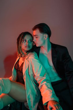 confident and stylish man in black blazer sitting near african american woman with dreadlocks wearing lace bodysuit and beige trench coat on grey background with red lighting clipart