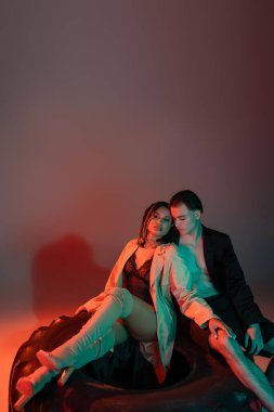 trendy interracial couple sitting on huge tire, shirtless man in black blazer and african american woman in lace bodysuit, beige trench coat and over knee boots on grey background with red lighting clipart