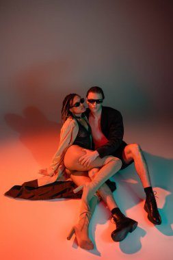 high angle view of trendy man in black blazer and shorts near african american woman in dark sunglasses, lace bodysuit, beige trench coat and over knee boots on grey background with red lighting clipart