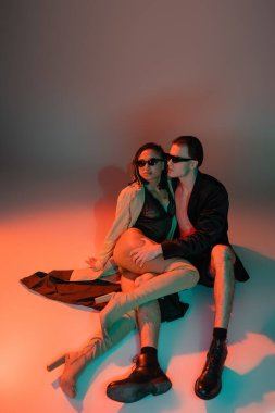 full length of glamour man in dark sunglasses and black blazer hugging leg of sassy african american woman in lace bodysuit, beige trench coat and over knee boots on grey with red lighting clipart