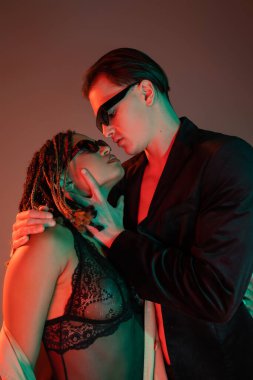 young and self-assured man in dark sunglasses and black blazer seducing enchanting african american woman in lace bodysuit on grey background with red lighting clipart