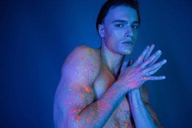 Karizmatik ve kendinden emin, ışıl ışıl ve çok renkli neon vücut boyalı tişörtsüz bir adam birleşik ellerle duruyor ve mavi arka plandaki Cyan ışık efektli kameraya bakıyor.