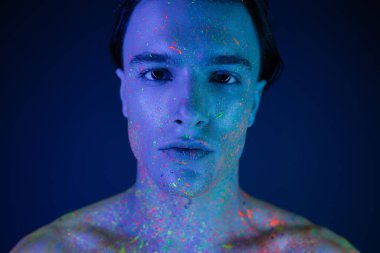 portrait of confident, self-assured man with bare shoulders posing in vibrant and colorful neon body paint while looking at camera on blue background with cyan lighting effect clipart