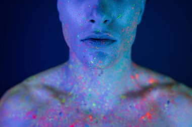 Genç ve gömleksiz bir adamın çok renkli ve neon renkli vücut boyası giymiş, bulanık mavi arka planda, siyan ışıklandırma efektiyle duran ve poz veren görüntüsü.