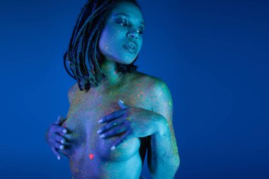 charming and nude african american woman posing in radiant and colorful neon body paint while looking away and covering breast with hands on blue background with cyan lighting effect clipart