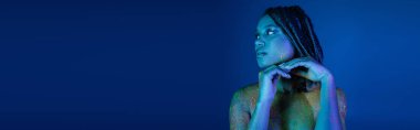 sexy and eye-catching african american woman with dreadlocks, in multicolored neon body paint, holding hands near chin and looking away on blue background with cyan lighting effect, banner